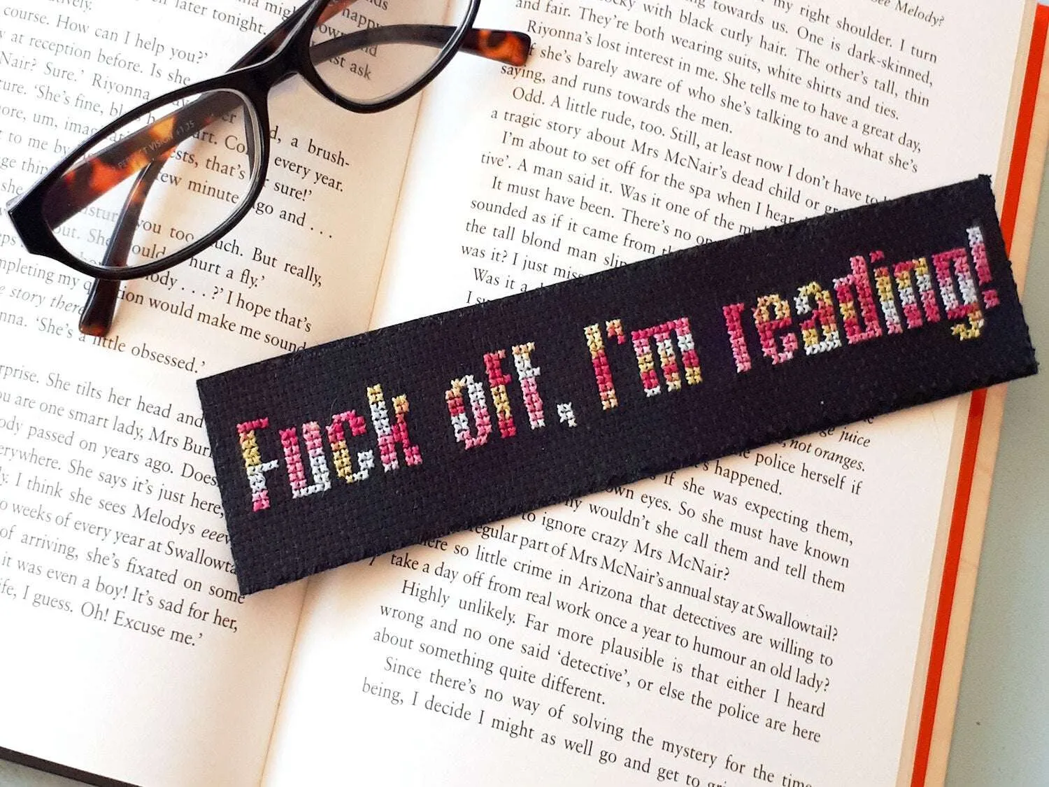 A black fabric bookmark and a pair of glasses lie over an open book. The bookmark is cross stitched with the words 'Fuck off, I'm reading!'