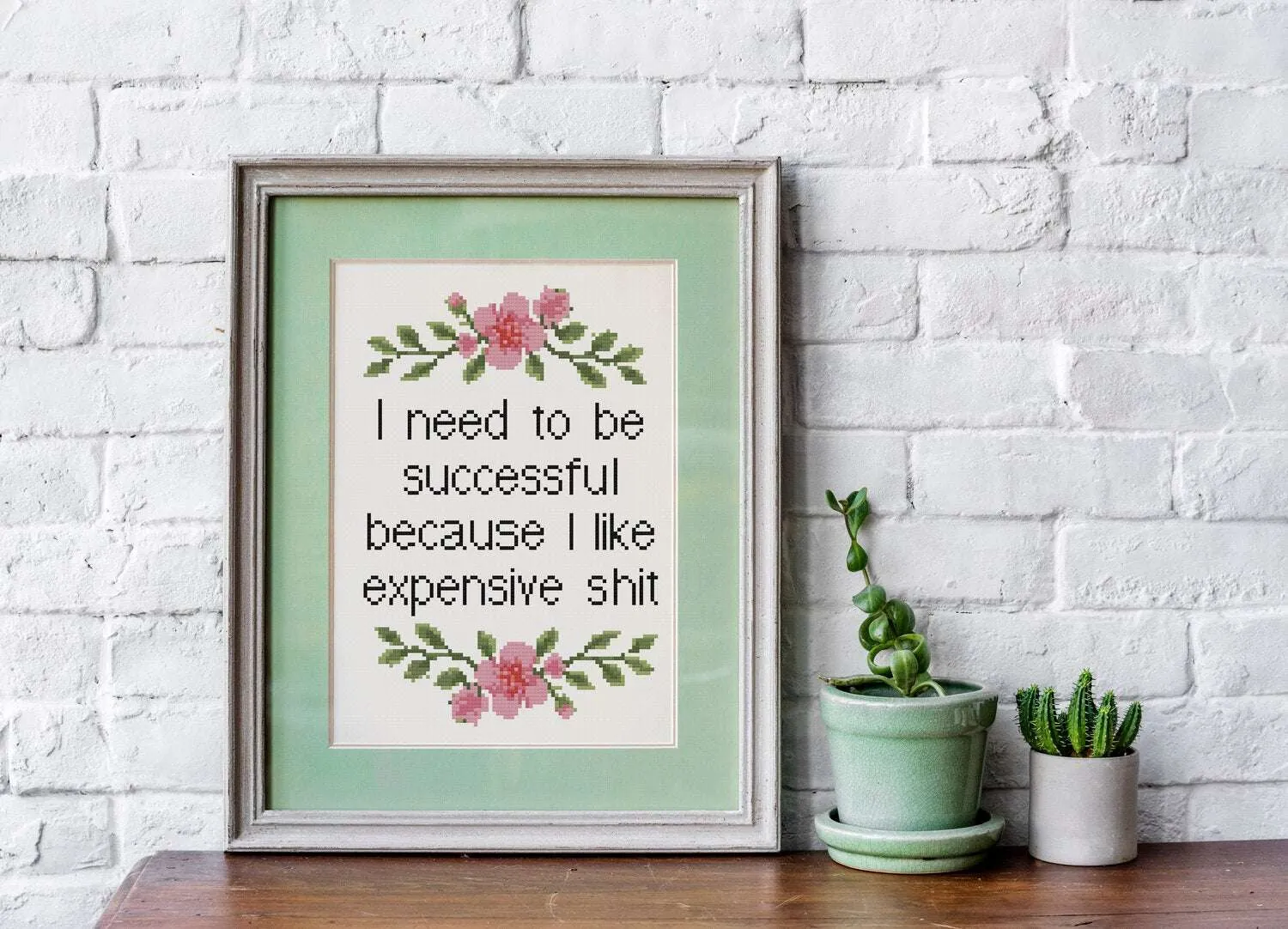 Framed cross stitch reading 'I need to be successful because I like expensive shit' in black with floral designs above and below