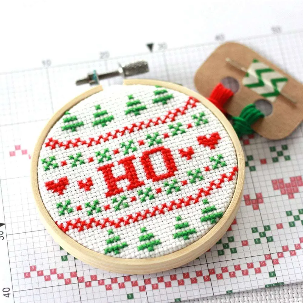 A cross stitch reading 'HO' in red text surrounded by red and green Christmas patterns, sits on a printed cross stitch pattern and next to some thread