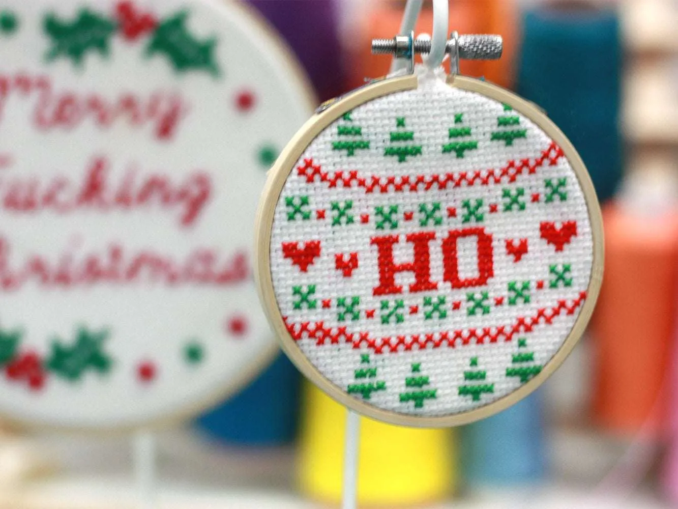 A cross stitch reading 'HO' in red text surrounded by red and green Christmas patterns, hangs in front of other cross stitch hoops