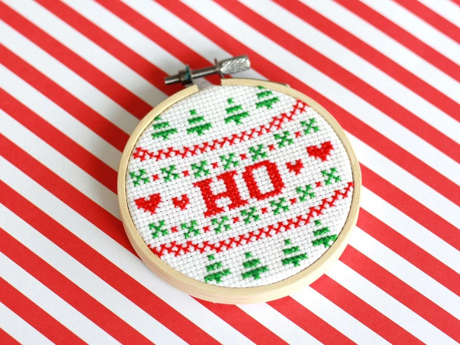 A cross stitch reading 'HO' in red text surrounded by red and green Christmas patterns, sits on a red and white striped background