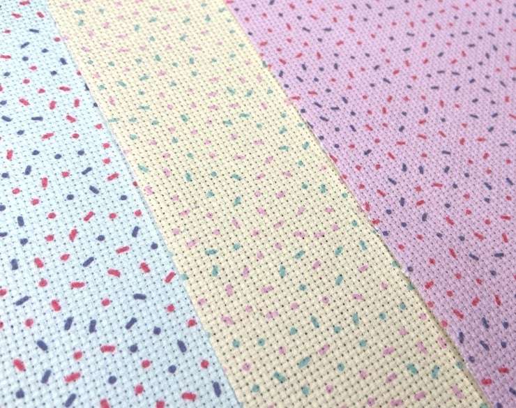 Three sheets of aida cross stitch lay flat, each patterned with donut sprinkles: one  pink, one mint green and one lemon yellow