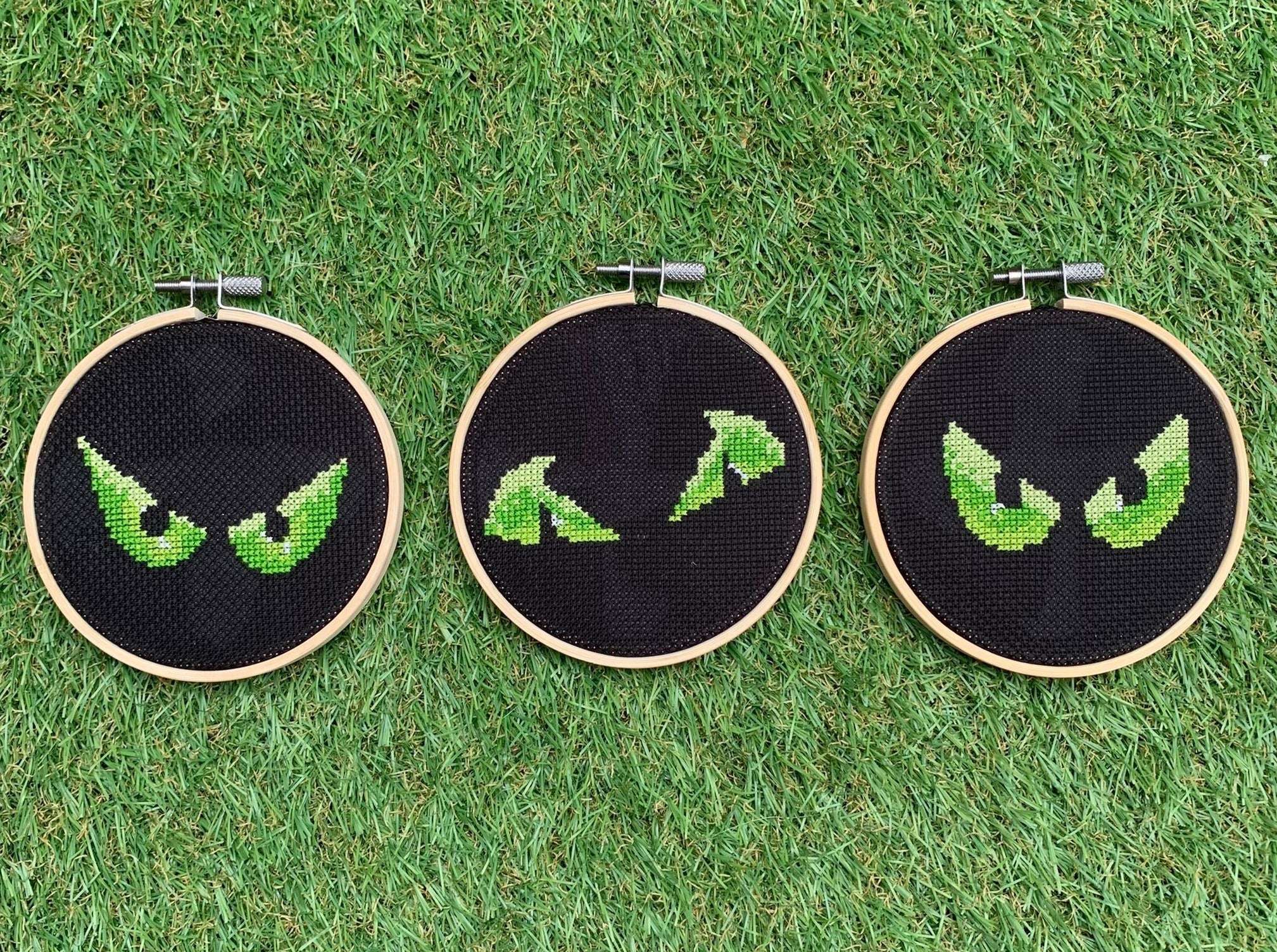 Three pairs of spooky eyes that are cross stitched in bright green onto black fabric