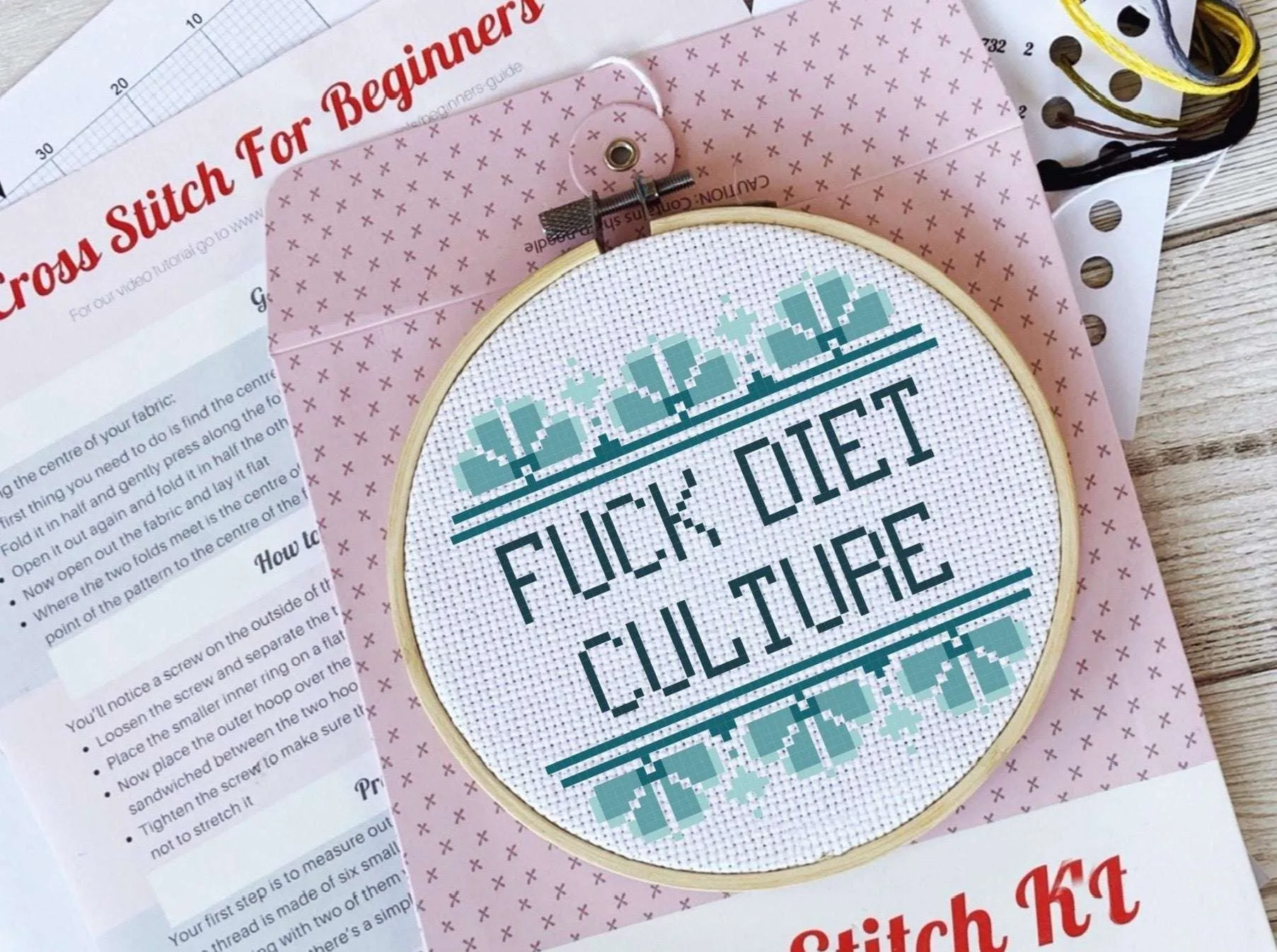 A framed cross stitch reads 'FUCK DIET CULTURE' surrounded by blue art deco patterns.