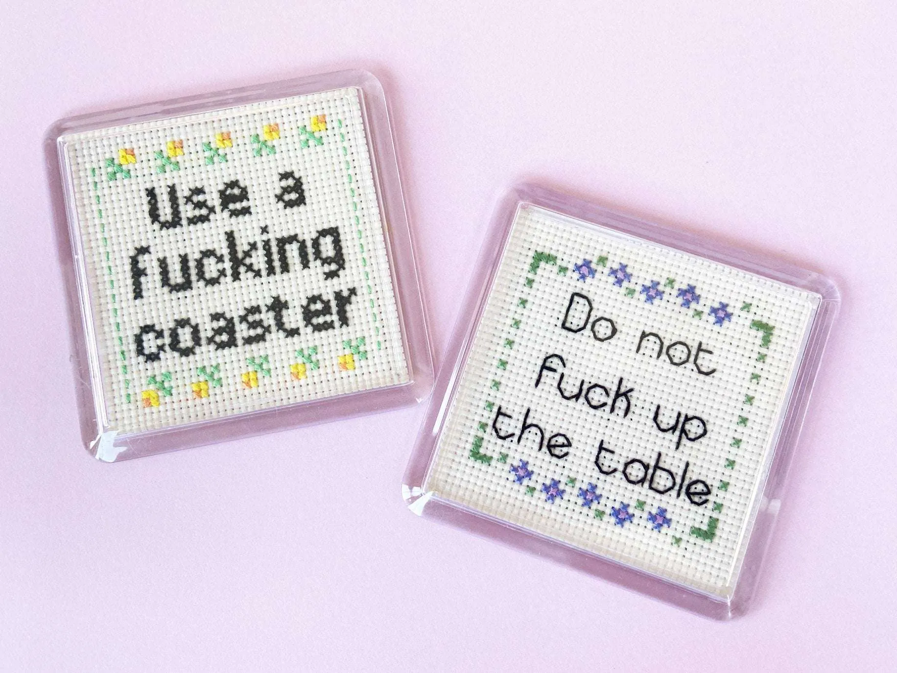 two clear plastic coasters with cross stitch inside. one coaster says use a fucking coaster and the other says dont fuck up the table