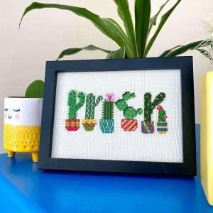 A framed cross stitch which features potted cacti that are shaped to spell the word prick