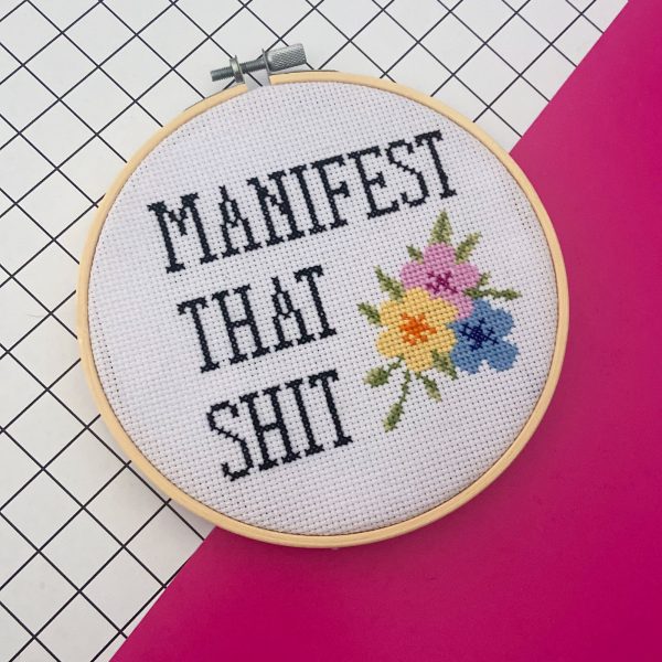 a cross stitched picture saying manifest that shit on a pink and white surface