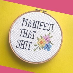 A cross stitch displayed in an embroidery hoop with the words manifest that shit. There are also some pretty flowers next to the writing