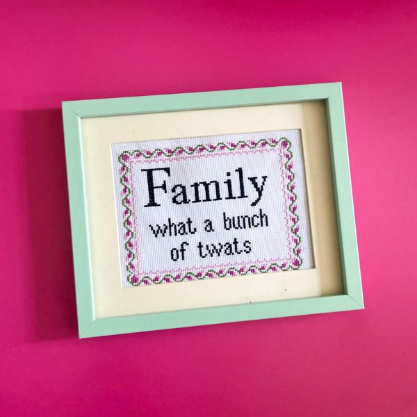 a framed cross stitch with the phrase “family, what a bunch of twats” on a pink wall